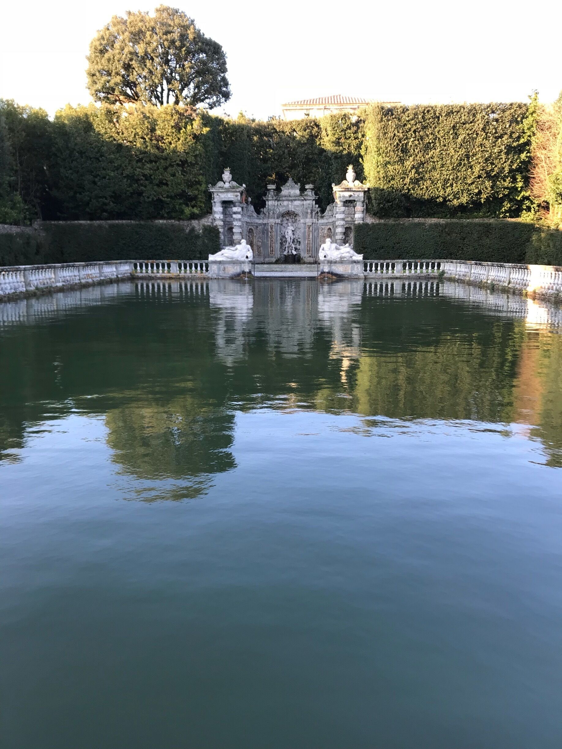 A serene water feature with statues and a fountain, surrounded by neatly trimmed hedges and trees.