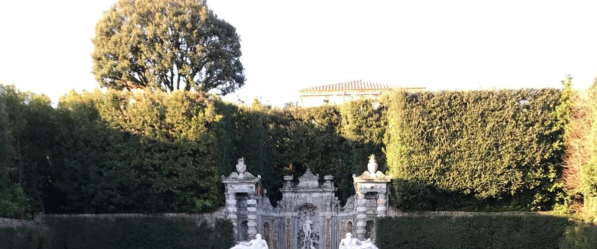 A serene water feature with statues and a fountain, surrounded by neatly trimmed hedges and trees.