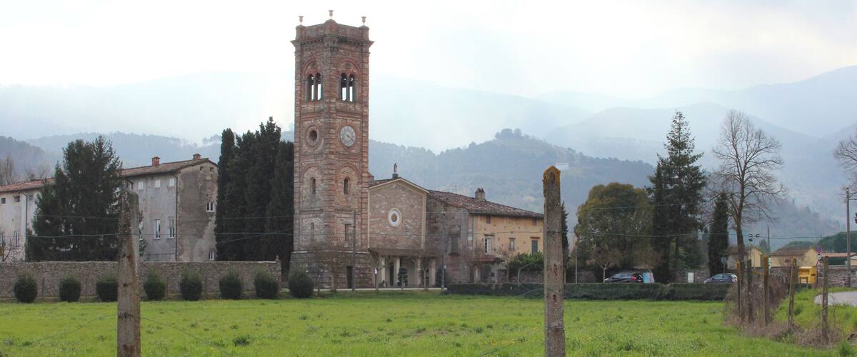 A historic church with a tall bell tower, surrounded by a grassy field and mountains in the background.