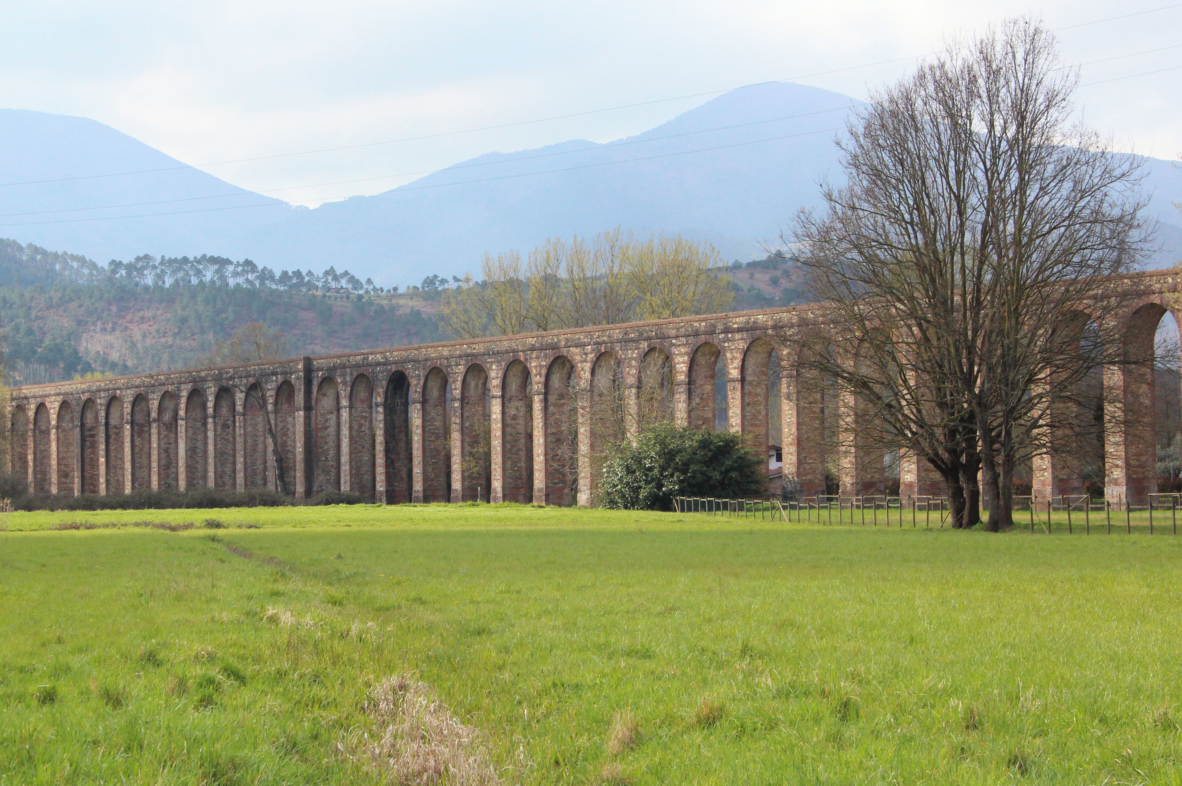A long brick viaduct with multiple arches, a grassy field in the foreground, and mountains in the background.