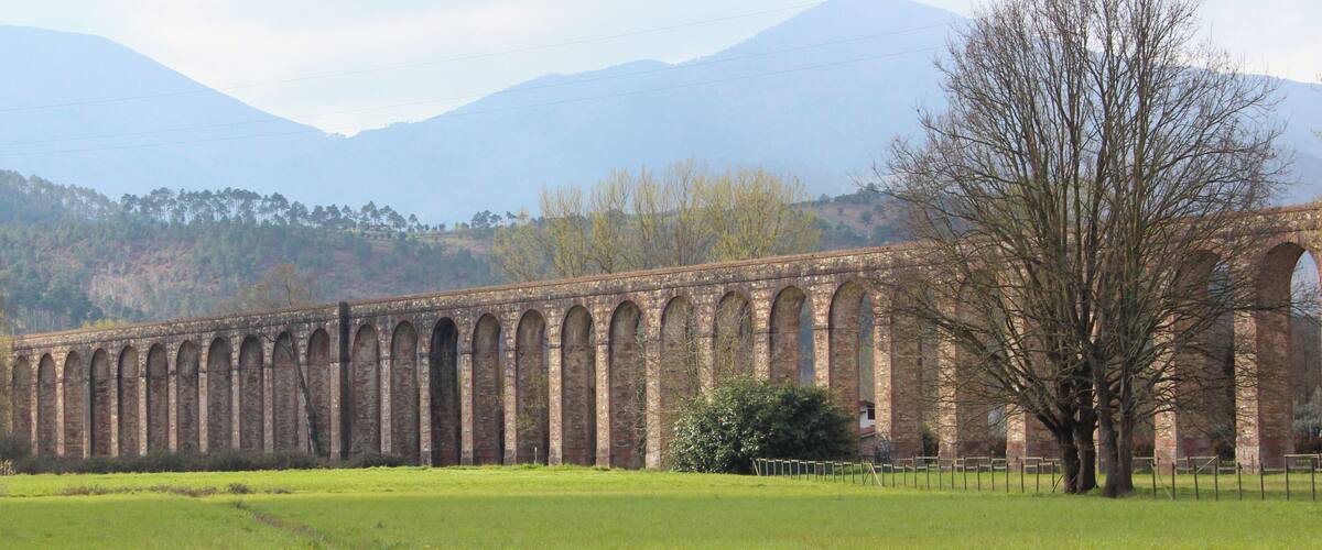 A long brick viaduct with multiple arches, a grassy field in the foreground, and mountains in the background.