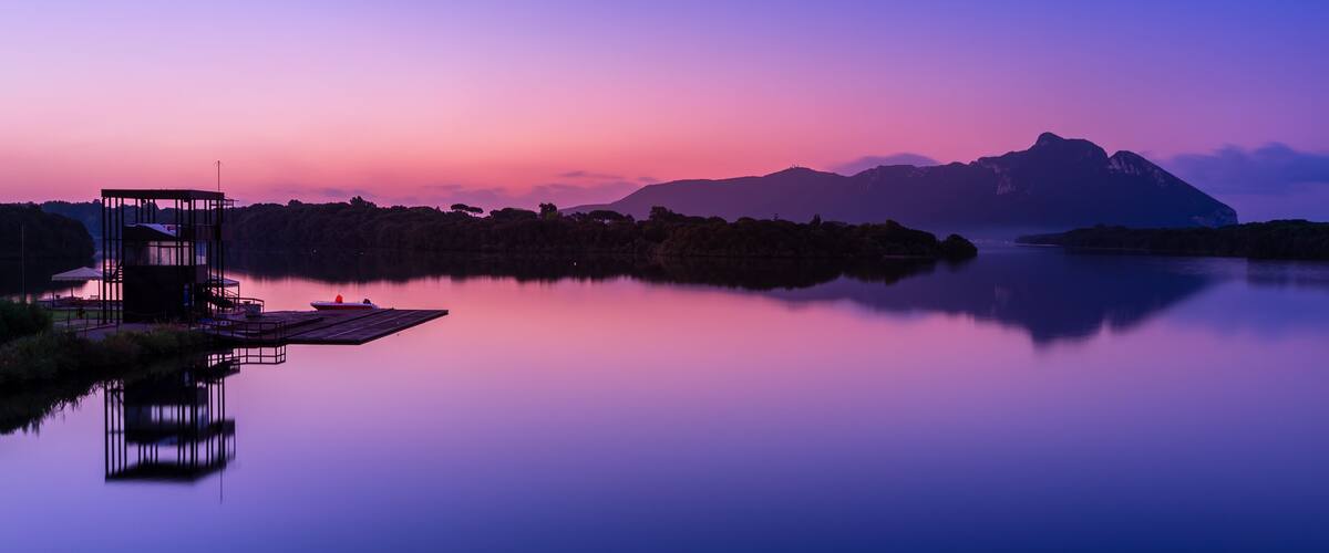 A serene lake at dusk with a dock and a small boat, surrounded by mountains and trees.