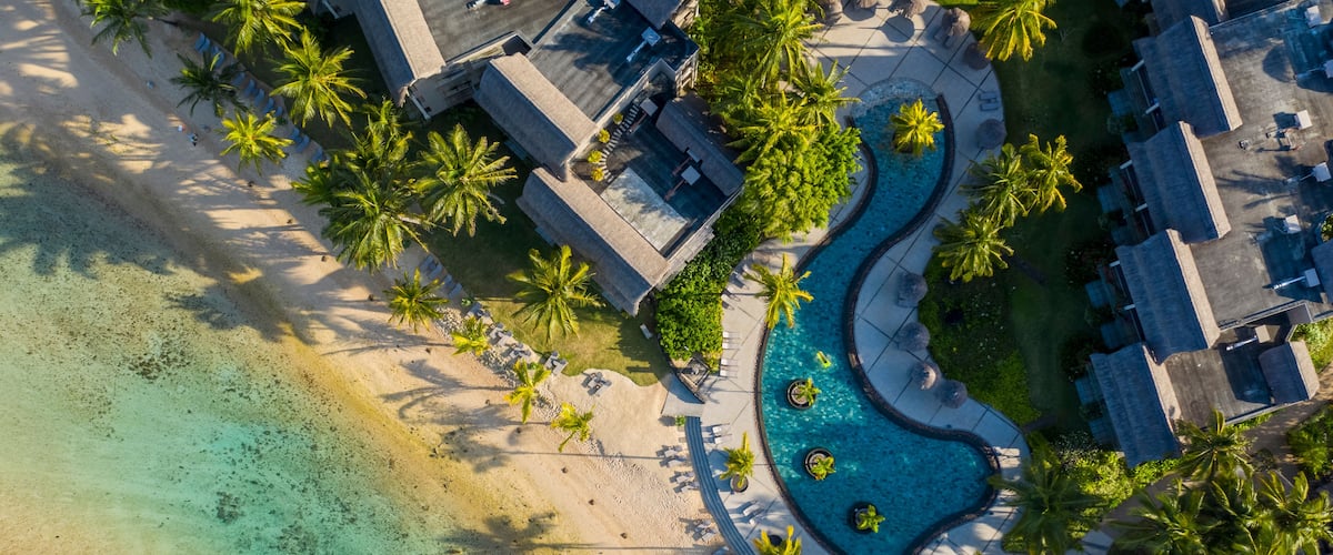 An aerial view of a resort with a swimming pool, palm trees, and a beach.