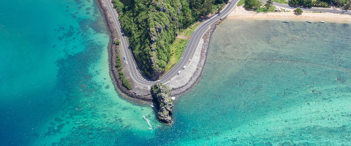 A coastal road curving through a forested area with clear turquoise waters and a rocky outcrop.