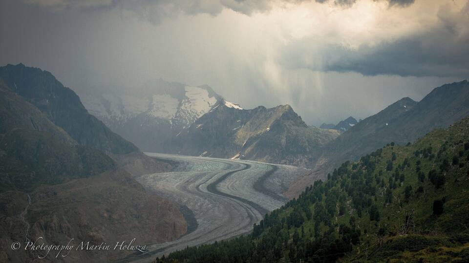 A glacier winding through a mountainous valley with snow-capped peaks and dense forests.