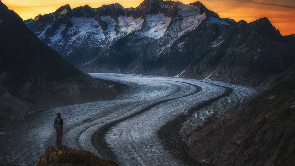 A glacier winding through a mountainous landscape with a person standing on a rocky outcrop.