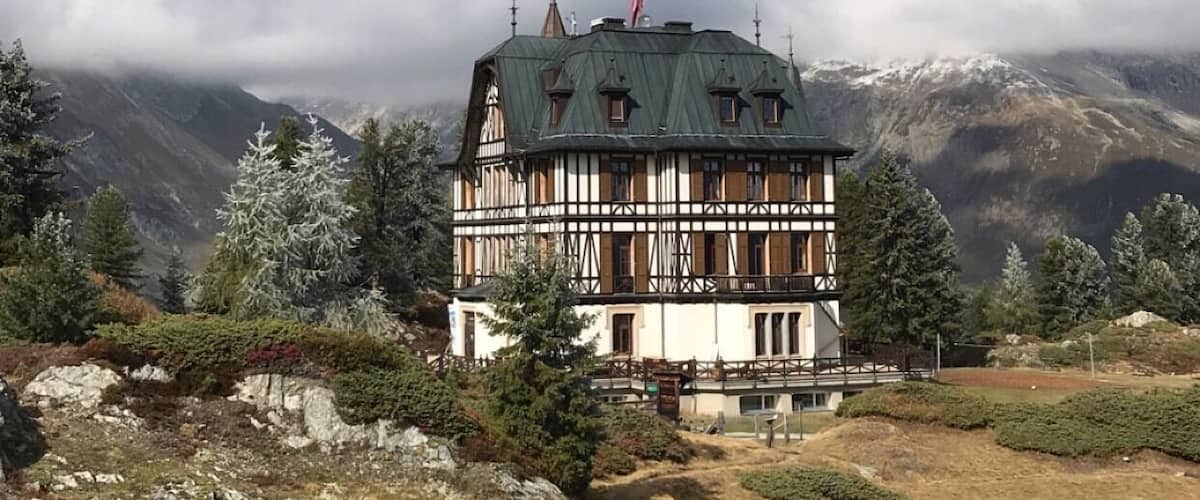 A traditional chalet with a steep roof, surrounded by mountains and snow-capped peaks.