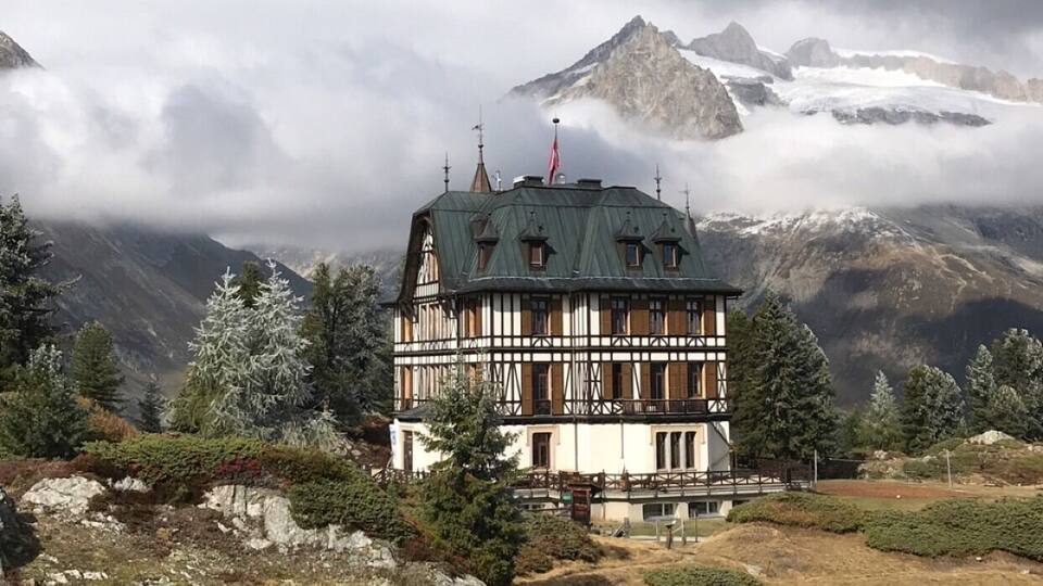 A traditional chalet with a steep roof, surrounded by mountains and snow-capped peaks.