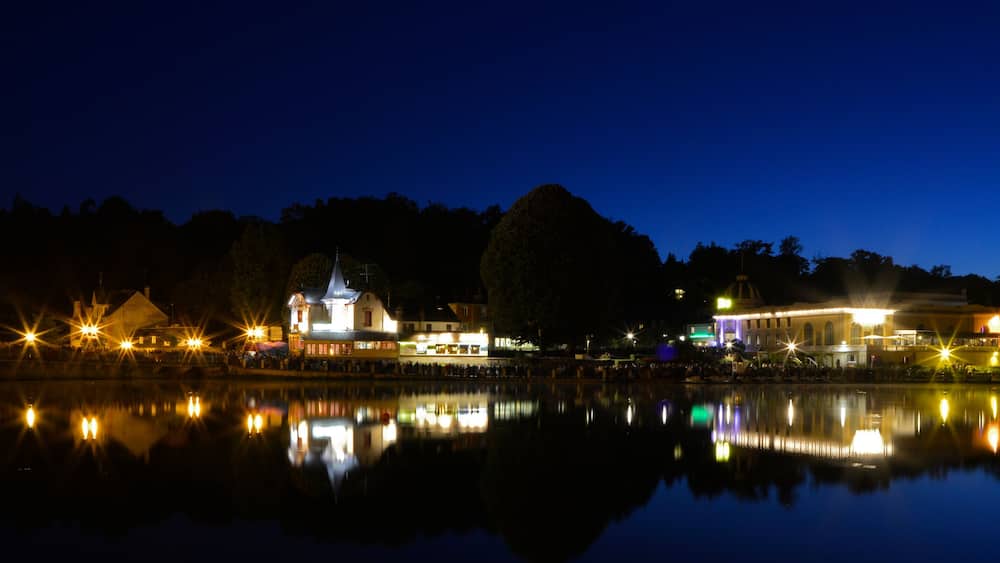 Une scène nocturne au bord du lac, avec des bâtiments et des arbres illuminés qui se reflètent dans l’eau.