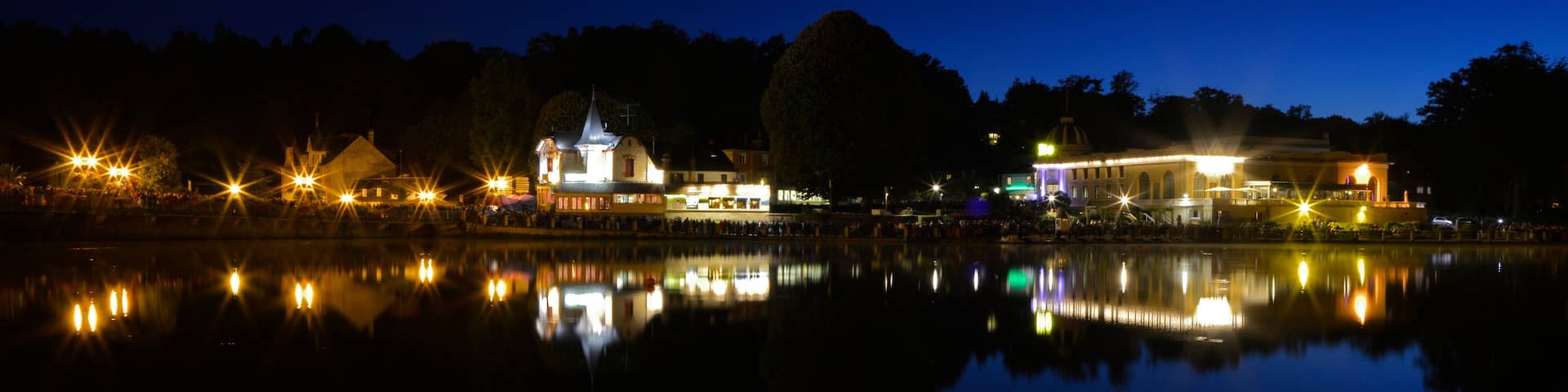 A nighttime lakeside scene with illuminated buildings and trees reflecting on the water.