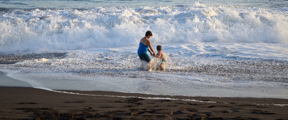 A beach with waves, a person and a child playing in the water, and a clear sky.