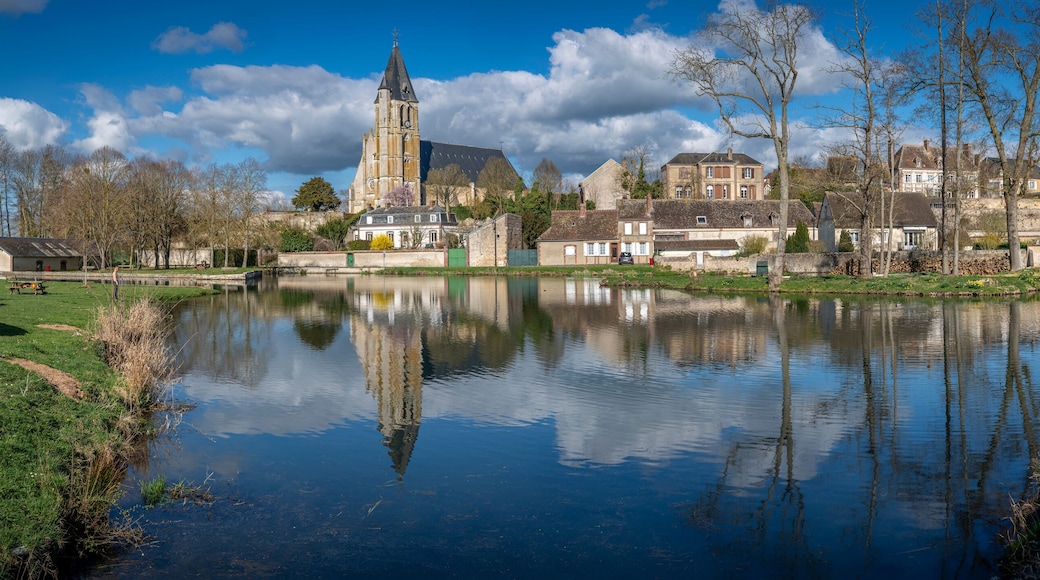Brezolles, France - 03 26 2025: Panoramic view of the rural villagewith Reflection of Saint-Nicolas church, trees, cloudy and blue sky on the pond water.