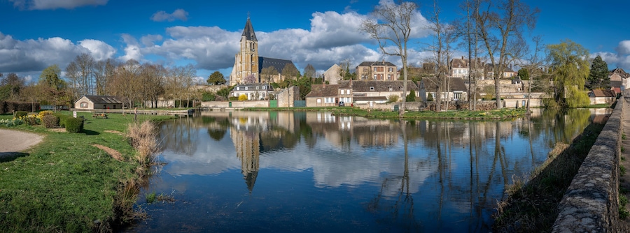 Brezolles, France - 03 26 2025: Panoramic view of the rural villagewith Reflection of Saint-Nicolas church, trees, cloudy and blue sky on the pond water.