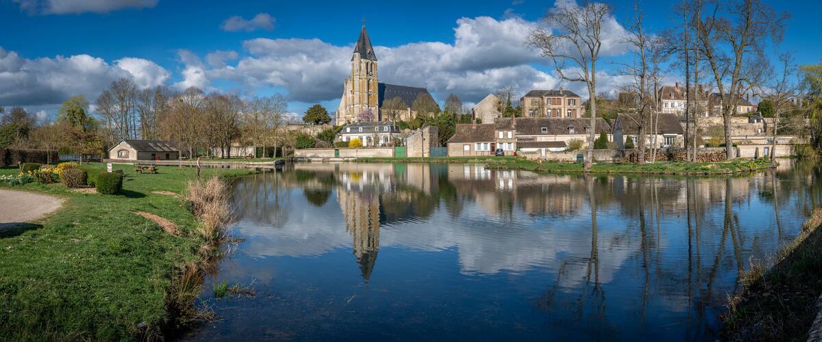 A calm river reflecting buildings and trees, with a church tower and residential houses along the banks.