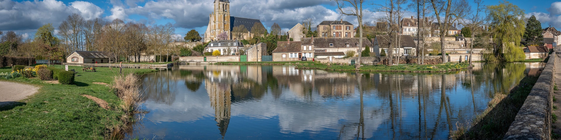 A calm river reflecting buildings and trees, with a church tower and residential houses along the banks.