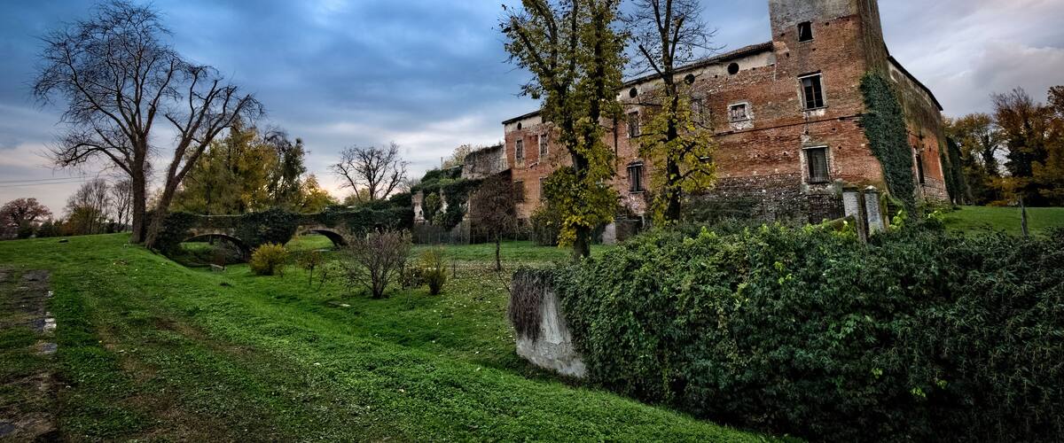 A historic brick building with a tower, surrounded by lush greenery and a stone bridge.