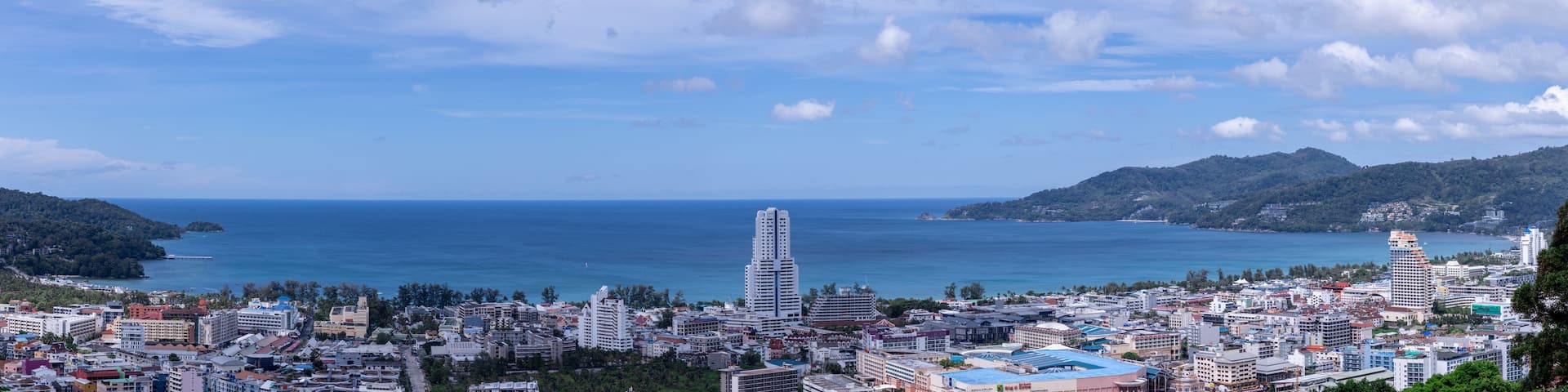 A coastal cityscape with a prominent white skyscraper, dense urban buildings, and a backdrop of hills and the sea.