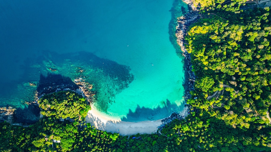 A coastal landscape with clear turquoise waters, a sandy beach, and dense green vegetation.