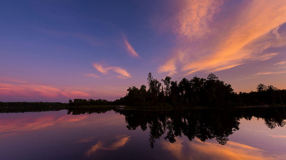 A calm lake reflecting a sunset sky with trees silhouetted on the opposite shore.