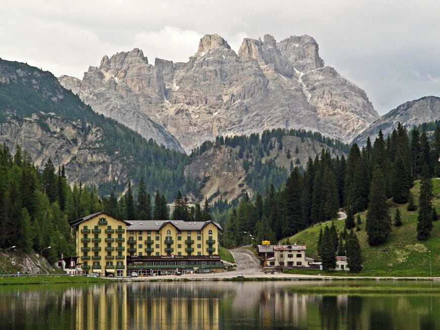 A mountain lodge with a lake in front, surrounded by forest and rocky peaks.
