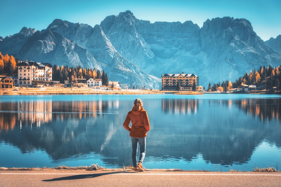 Young woman standing on the coast of Misurina lake at sunrise in autumn. Dolomites, Italy. Landscape with girl in red jacket, reflection in water, buildings, blue sky. Italian alps. Travel background