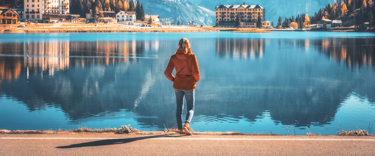 A person standing by a calm lake with mountains in the background.