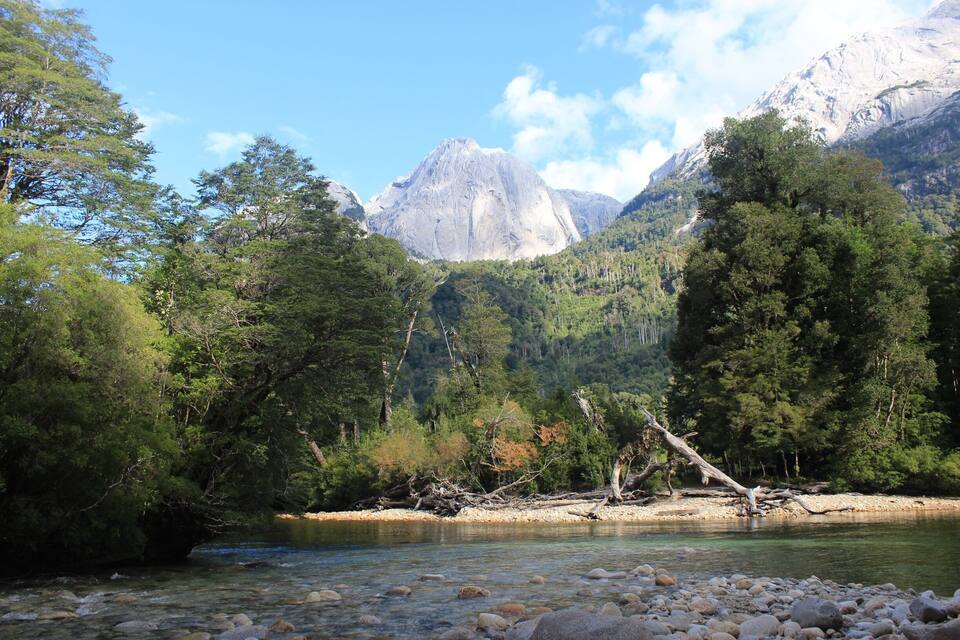 A river with clear water, surrounded by dense green forest and rocky terrain, with a mountain in the background.