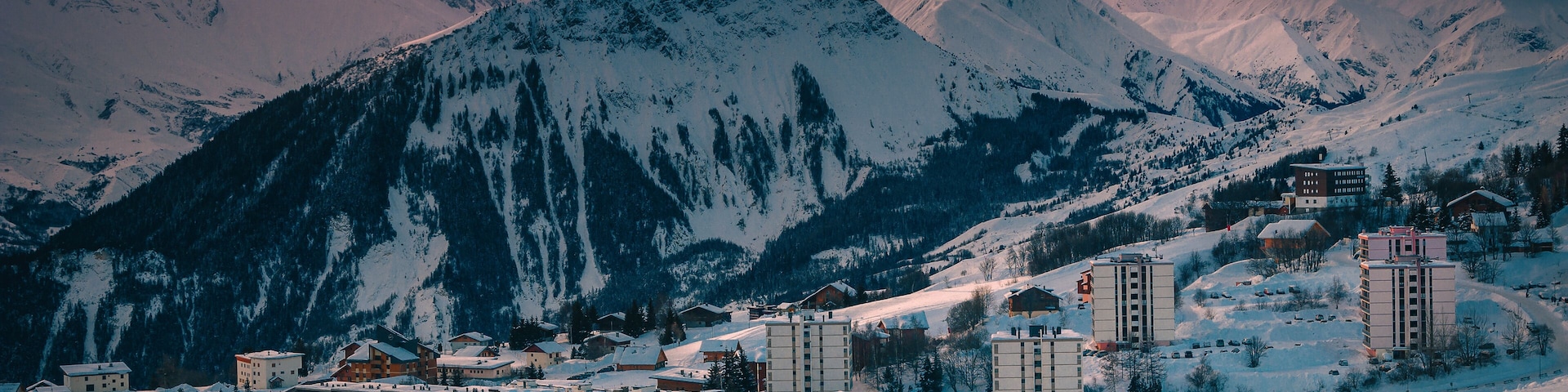 A snow-covered mountain range with a town at the base, featuring buildings and a ski slope.