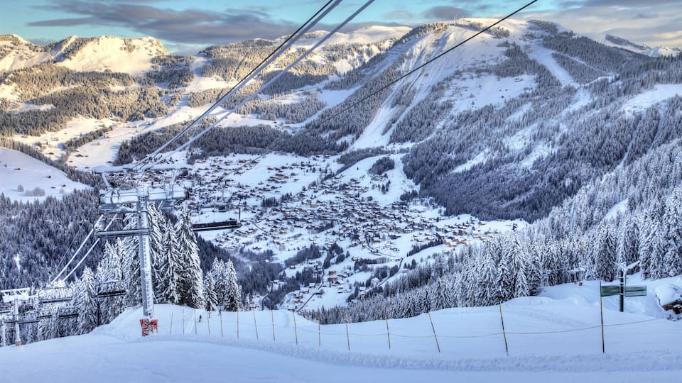 A snow-covered mountain village with ski lifts, surrounded by dense forests and distant peaks.