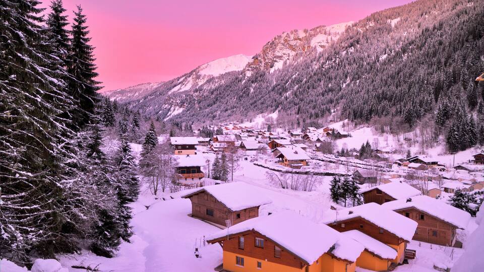A snow-covered village with wooden houses and dense evergreen trees.