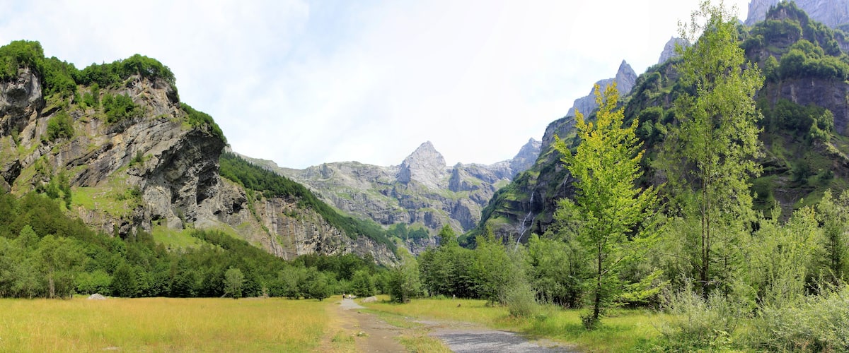 A mountainous landscape with a dirt path, dense green forest, and rocky cliffs.
