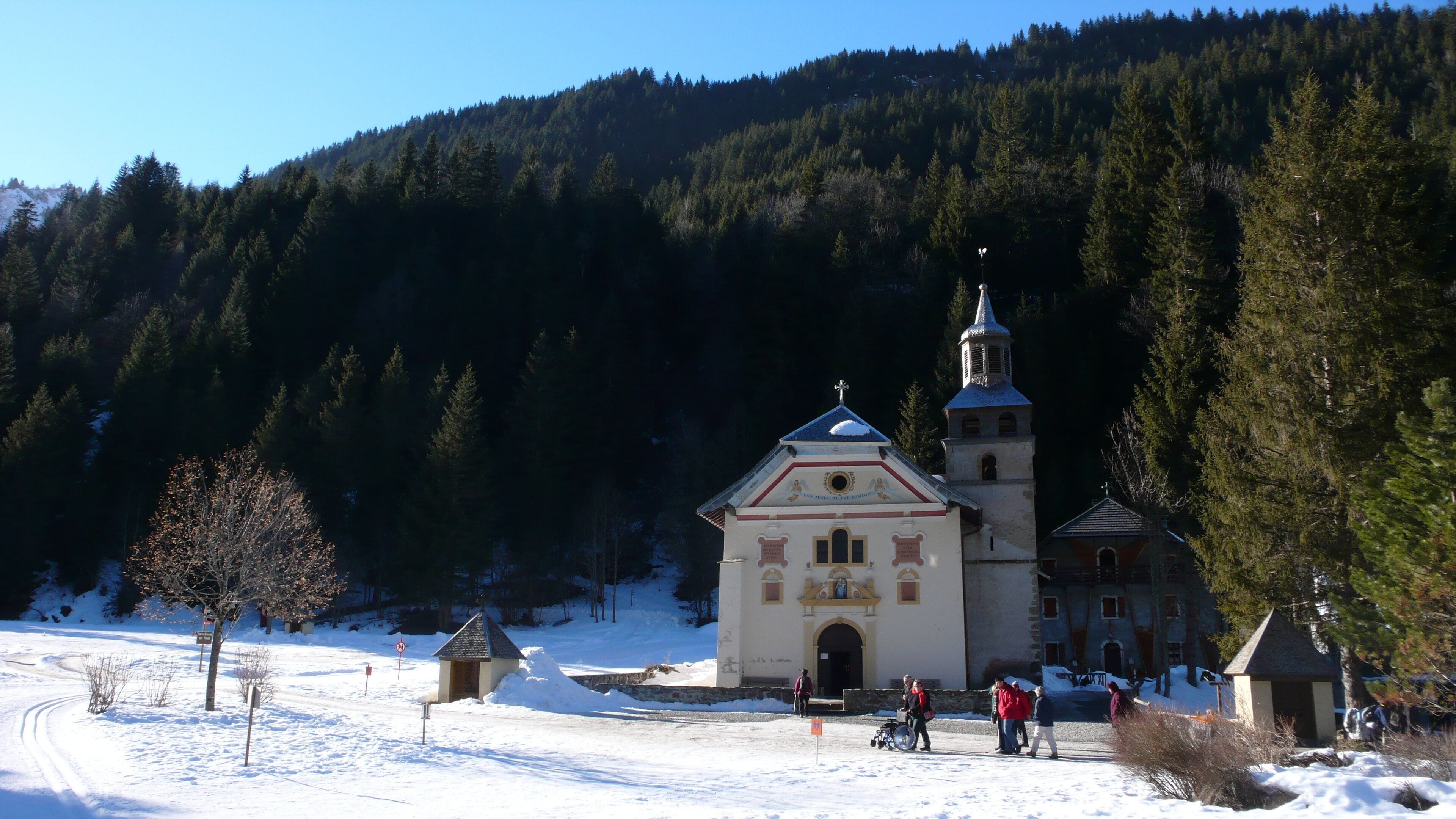 A snow-covered church with a bell tower, surrounded by a forested mountain.