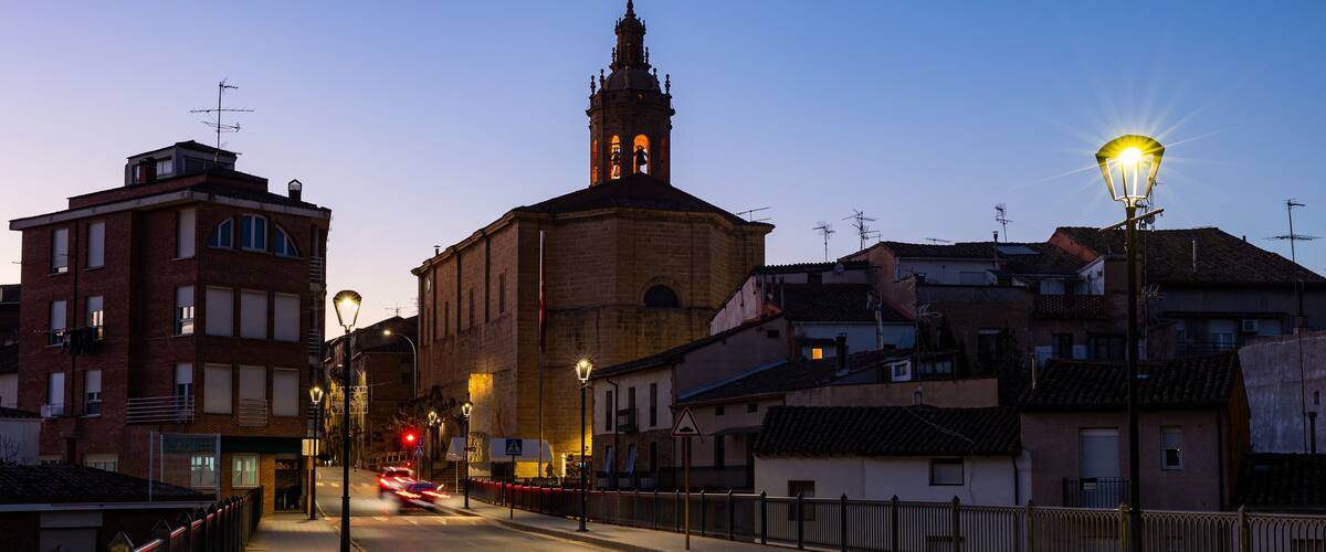 A street at dusk with a prominent church tower, street lamps, and residential buildings.