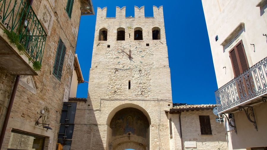 Une haute tour en pierre, dotée de fenêtres en arc de cercle et d’une horloge, entourée d’immeubles avec des balcons et des volets.