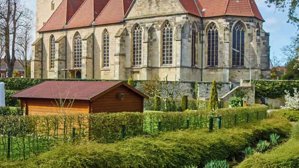 A historic church with a red-tiled roof and stone walls, surrounded by well-maintained gardens and a small stream.