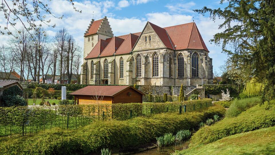 A historic church with a red-tiled roof and stone walls, surrounded by lush greenery and a small stream.