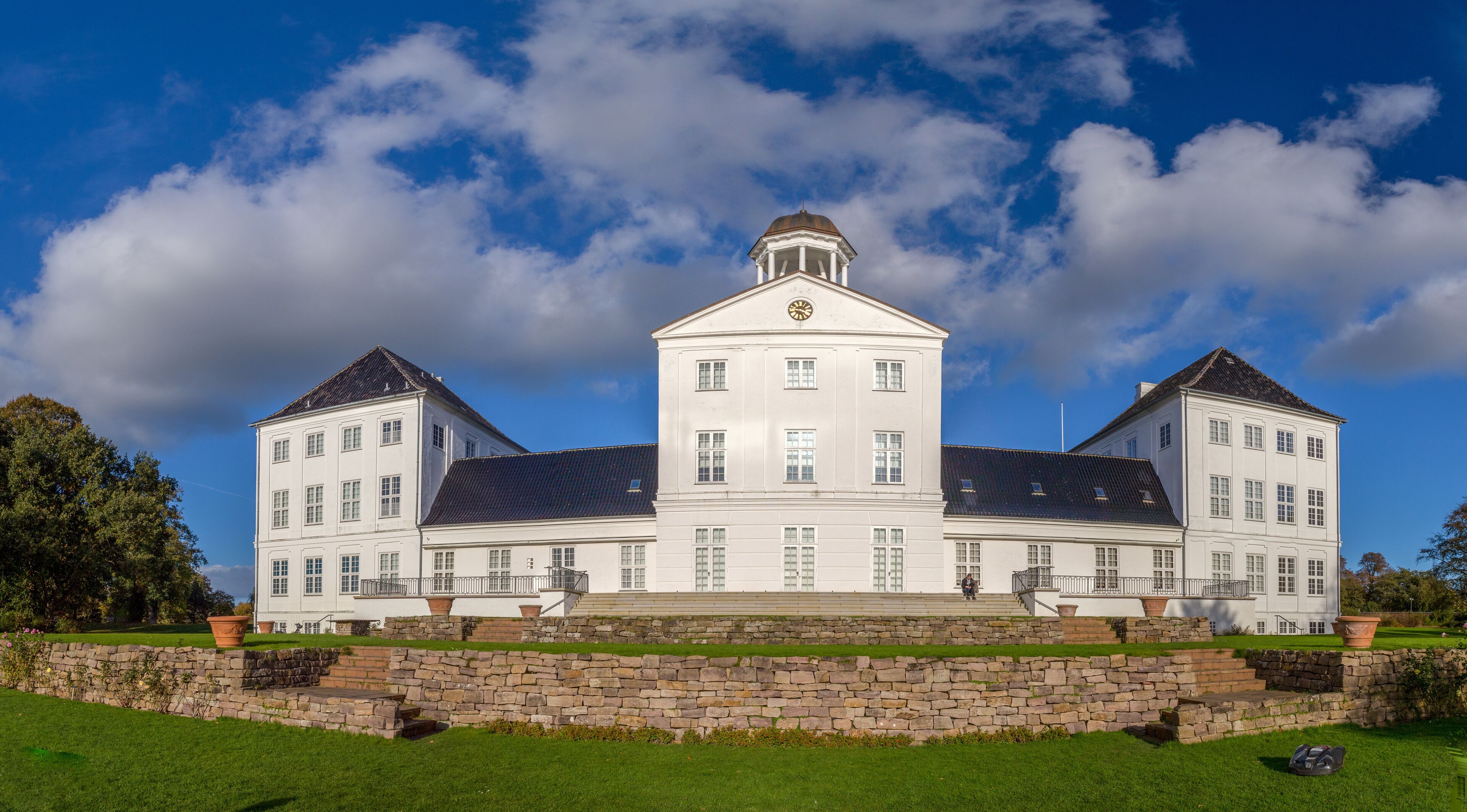 A large white building with a central tower and two symmetrical wings, surrounded by a stone wall and green lawn.
