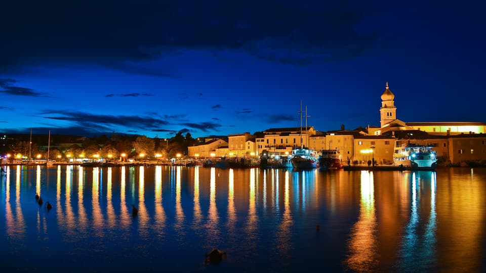 A nighttime waterfront with illuminated buildings and a prominent tower reflecting on the water.
