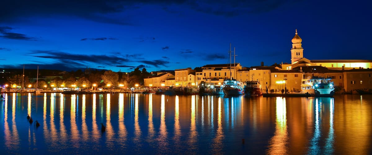 A nighttime waterfront with illuminated buildings and a prominent tower reflecting on the water.