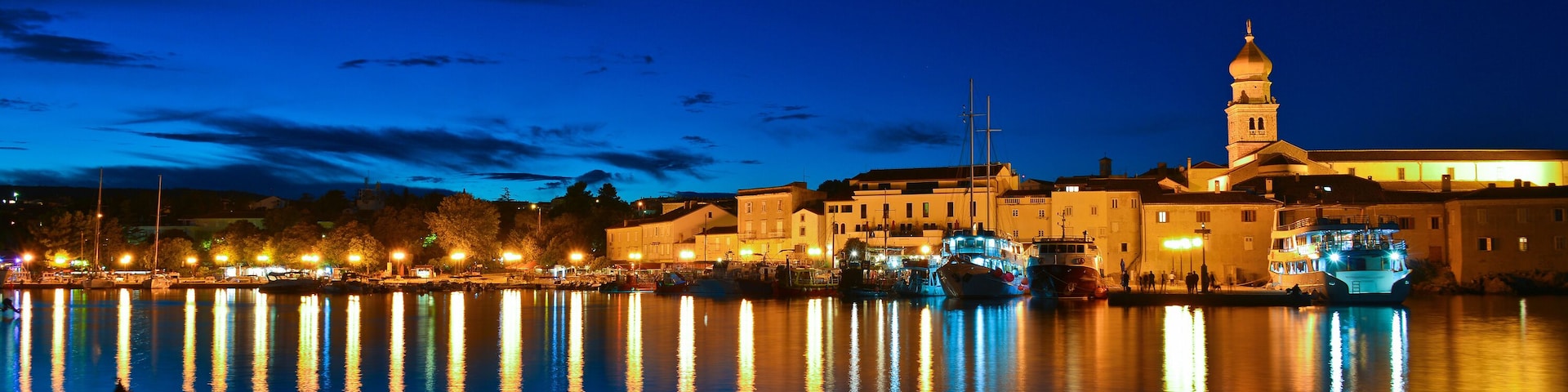 A nighttime waterfront with illuminated buildings and a prominent tower reflecting on the water.