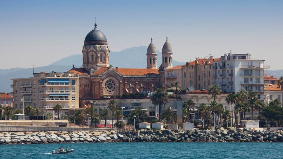 A coastal cityscape with a prominent cathedral featuring two domes, a casino, and a rocky shoreline with a small boat.