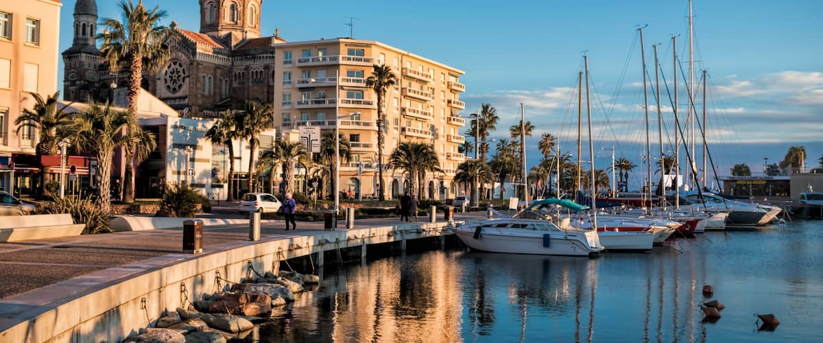 A marina with boats, a historic building with a dome, palm trees, and a clear sky.