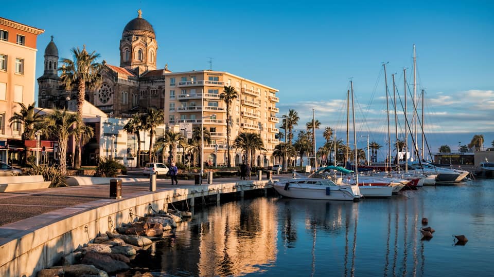 A marina with boats, a historic building with a dome, palm trees, and a clear sky.