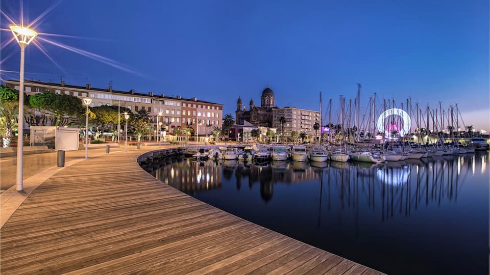 A waterfront promenade with a wooden boardwalk, boats docked in the marina, and a prominent church with a dome.