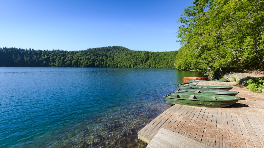 Un lac aux eaux limpides, où des bateaux sont amarrés à un quai en bois, le tout entouré d’une forêt dense.