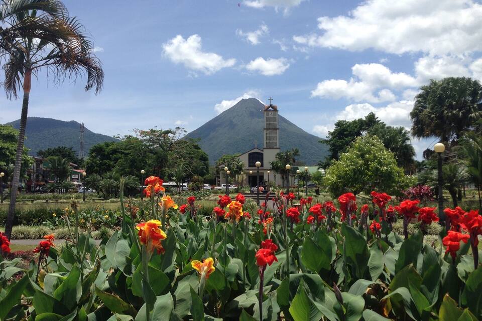 Eine Kirche mit einem Glockenturm, umgeben von einem Garten mit roten und orangefarbenen Blumen, Palmen und einem Berg im Hintergrund.