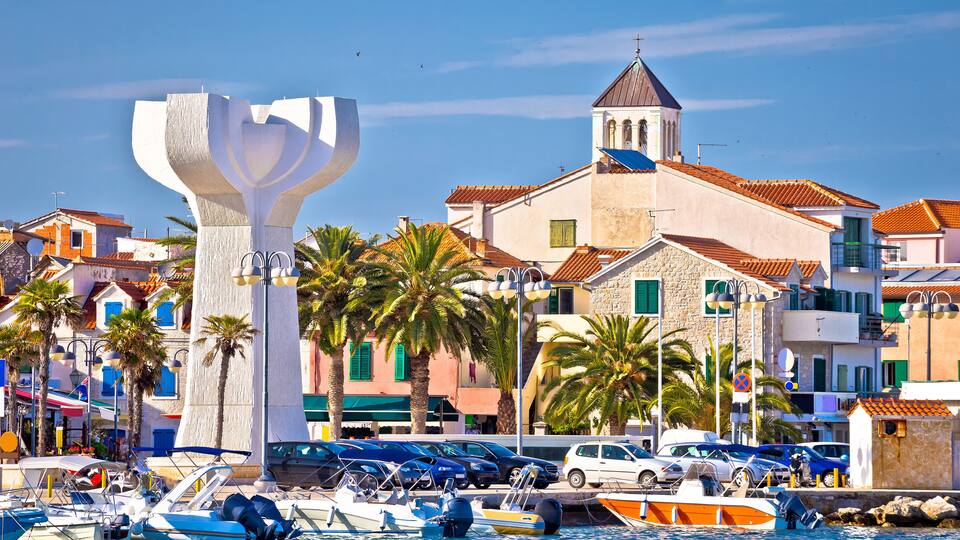 A coastal town with a prominent white sculpture, parked cars, and a church with a bell tower.