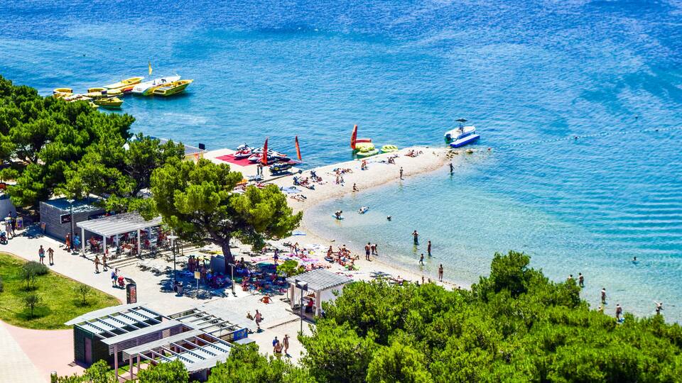 A coastal area with a beach, boats, and a pier with people and umbrellas.