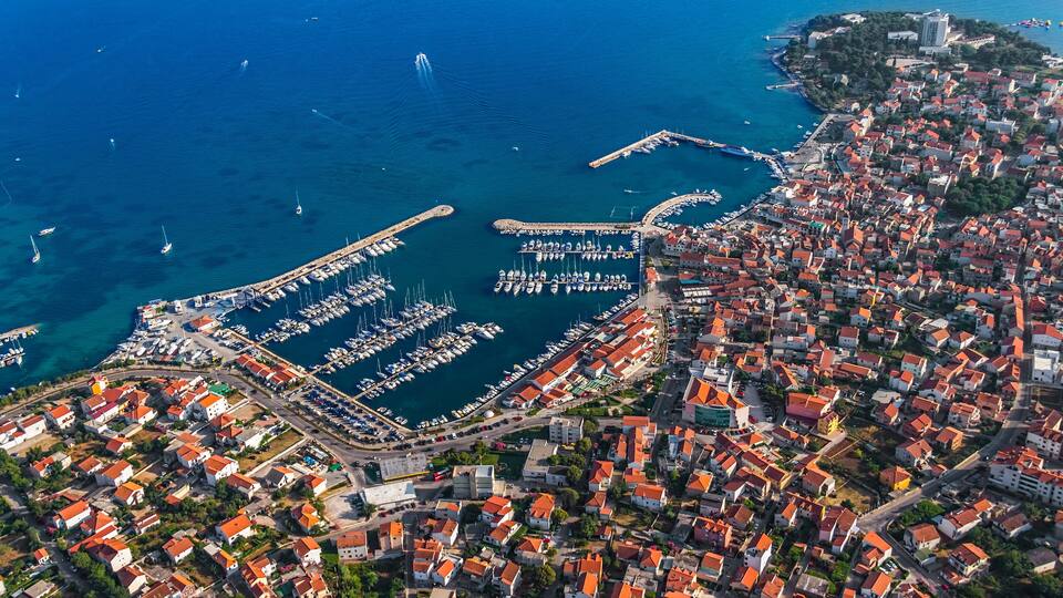 An aerial view of a coastal town with a marina, red-roofed buildings, and a clear blue sea.