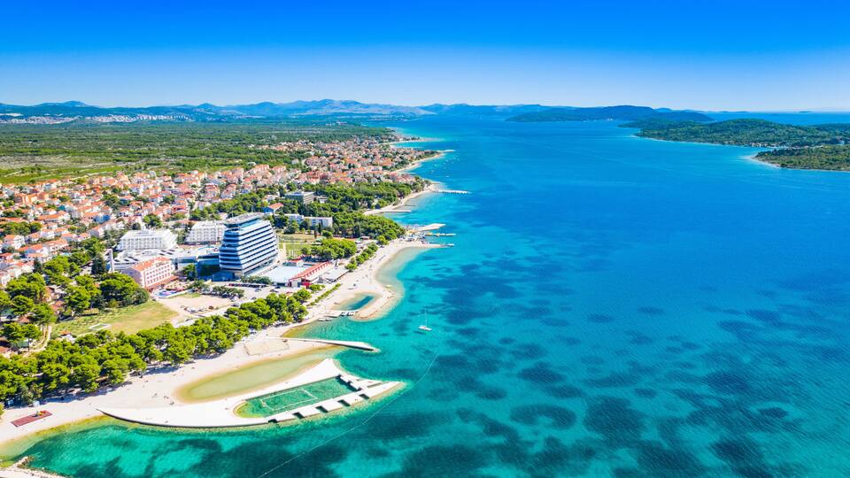 An aerial view of a coastal town with a clear blue sea, a sandy beach, and a mix of residential and commercial buildings.